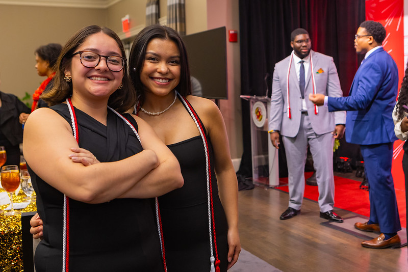 2 IAP students wearing graduation cords and stage in background